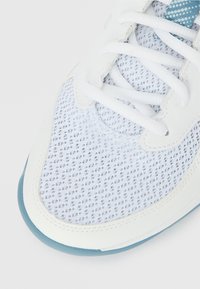 Κοντινό πλάνο στην μύτη λευκού αθλητικού παπουτσιού με πλεκτό ύφασμα, λευκά κορδόνια και λεπτομέρεια στη σόλα σε ανοιχτό μπλε χρώμα σε απλό φόντο.