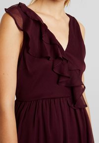Burgundy klänning med volangärmar och V-ringning. Tyget verkar lätt och har en texturerad yta, med en samlad midja.