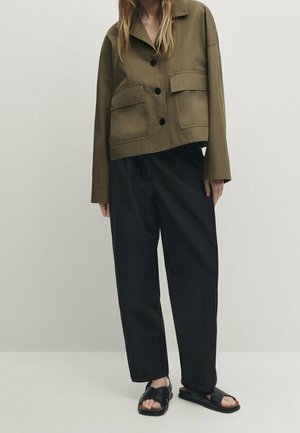 Femme portant une veste boutonnée vert olive avec de grandes poches, un pantalon noir ample et des sandales noires à bouts ouverts, debout devant un fond uni.