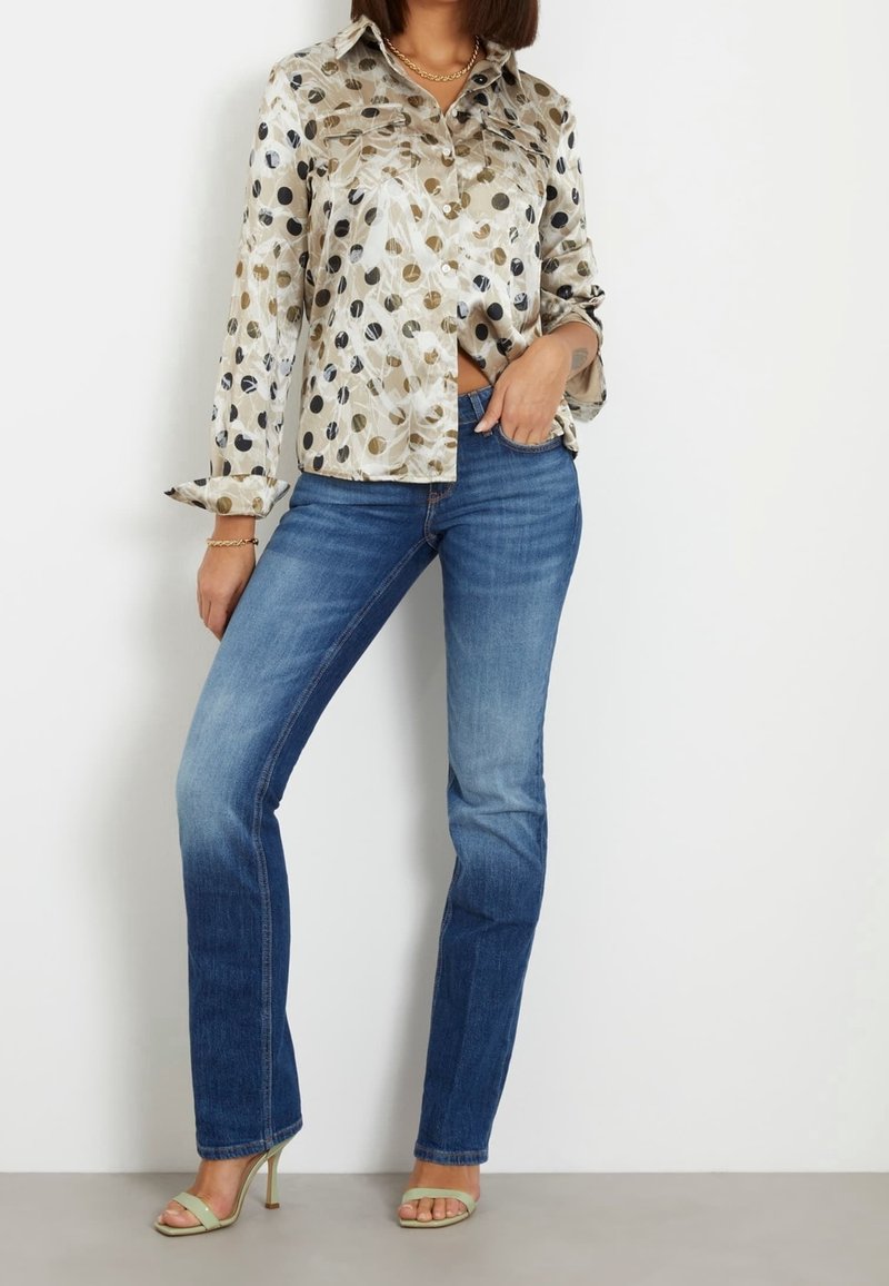 Chemise boutonnée à motifs pois beige et multicolores, associée à un jean en denim bleu et des sandales à talons vertes.