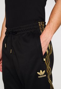 Svarta atletiska joggers med guld sidestreck, elastisk midja och dragsko. Har en sidficka och guld Adidas-logga.