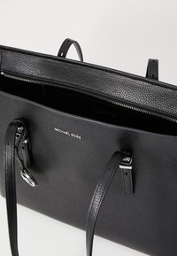 Black leather tote bag with a pebbled texture, silver hardware, and dual handles. Interior visible with zip closure and logo detail.