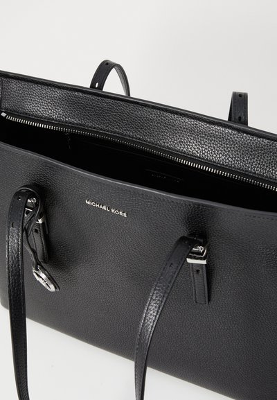 Sac fourre-tout en cuir noir avec une texture granuleuse, matériel en argent et poignées doubles. Intérieur visible avec fermeture éclair et détail du logo.
