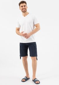 T-shirt bianco a V, pantaloni cargo blu navy con due tasche laterali, e ciabatte blu con testo bianco. Design e vestibilità casual e semplice.