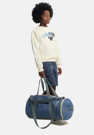 Niño con pelo rizado que lleva una sudadera clara y pantalones cortos azul marino, sosteniendo una bolsa de viaje azul con correas grises.