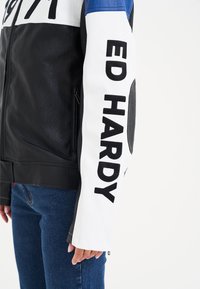 Giacca in pelle nera con accenti bianchi e blu, grande lettering nero sulla manica, tasca con zip e dettagli testurizzati sulle spalle.