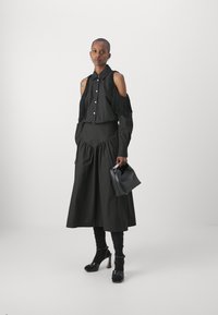 Camicia nera con spalle frangiate e bottoni, abbinata a una gonna nera a balze. Gli accessori includono una piccola borsa nera e scarpe con tacco alto.