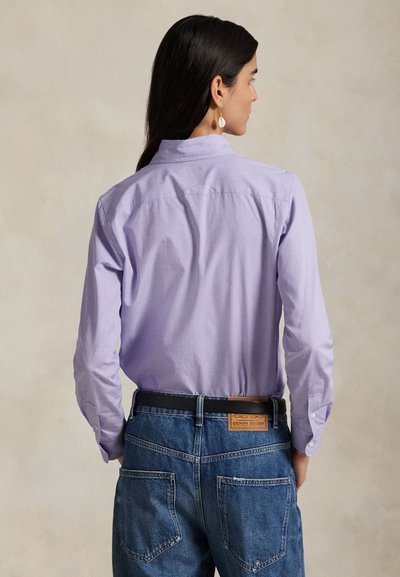 Pitkät tummaat hiukset omaava nainen pukeutuneena vaalean purppuraan pitkähihainen paita sinisten farkkujen kanssa, musta vyö vyötettynä, selin.