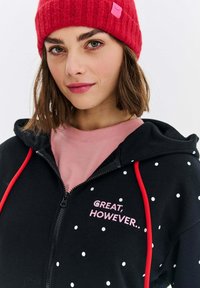 Czarna bluza z kropkami i czerwonymi sznurkami, z haftem "WSPANIALE, JEDNAK..." w różowym kolorze. Nosi na sobie różową bluzę. Czerwona czapka dzianinowa.