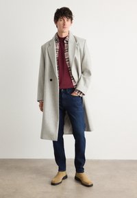 Light gray wool coat with a wide collar over a maroon sweater and plaid shirt. Dark jeans and tan ankle boots complete the outfit.