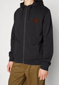 Persona che indossa una felpa nera con zip e un piccolo logo marrone, con pantaloni beige, in piedi contro uno sfondo chiaro e uniforme.