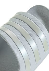 Gros plan d'une sandale légère grise avec trois bandes horizontales brillantes légèrement courbées sur la sangle. La sandale présente une surface texturée.