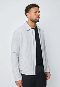 Homme avec des cheveux courts et bouclés et une barbe, portant une veste zippée grise claire sur une chemise noire, se tenant devant un fond clair uni.