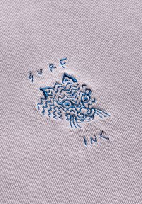 Wyhaftowany wzór na jasnoszarym materiale przedstawiający stylizowany wizerunek kota w niebieskiej nici, z napisami "SURF" i "INC" poniżej.