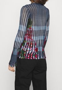 Transparent långärmad skjorta med vertikala blå och vita ränder, med blommiga dekorationer i rött och grönt längs fållen och ärmarna.