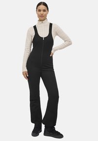 Sort biboverall med lynlås foran, tætsiddende design og bred ben. Underneath en lysebeige top med mock neck og tekstureret mønster. Sorte sneakers.