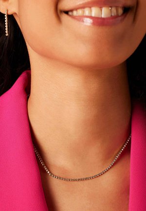 Collier en argent avec de petites pierres rondes et scintillantes, porté sur un décolleté lisse, contre un vêtement rose vif.