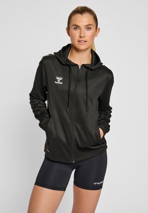 Jonge vrouw met blonde paardenstaart draagt een zwarte hoodie met rits en zwarte sportshorts, handen in de jaszakken, tegen een effen achtergrond.