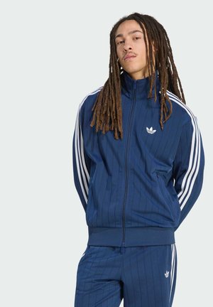 Młoda osoba z długimi dredami, ubrana w niebieski dres Adidas z białymi paskami, stoi z rękoma za plecami.