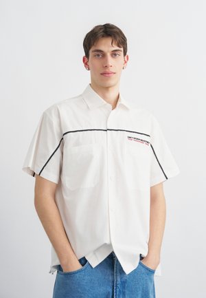 Joven con camisa blanca de manga corta y botones, con ribetes negros, y jeans azules, de pie con las manos en los bolsillos sobre un fondo liso.
