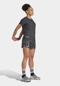 Sportoutfit bestehend aus einem enganliegenden, dunkel getarnten T-Shirt und passenden Shorts. Graue Turnschuhe mit weiß-gestreiften Socken in Schwarz.
