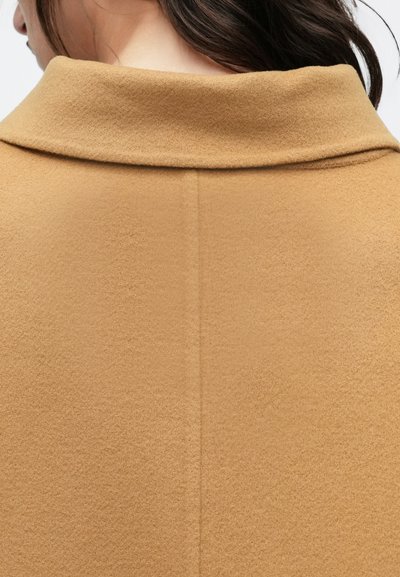 Gros plan sur le dos d’une personne portant un manteau en laine couleur camel avec un large col plié et une couture centrale.
