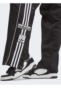 Svarta träningsbyxor med vit tre-stripes design, med tryckknappar och en Adidas-logotyp. Svarta och vita sneakers med mockadetaljer.