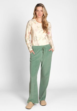 Woman standing with hands in pockets wearing floral long-sleeve top, green drawstring pants, and beige slippers on white background.