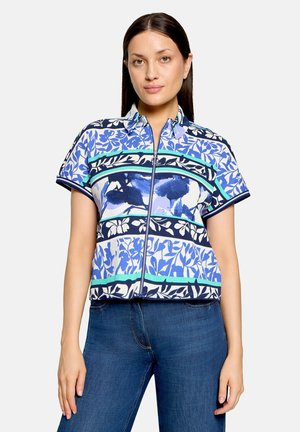 Femme portant un haut zippé à manches courtes avec des motifs floraux et abstraits bleu, blanc et vert, associé à un jean bleu.