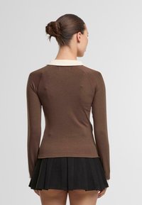 Ruskea pitkähihainen paita, jossa on kermahuppu, istuva muotoilu ja pehmeä, teksturoitu kangas. Yhdistetty mustaan, poimutettuun hameeseen.