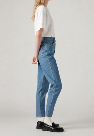Femme debout de profil, portant un t-shirt blanc, un jean bleu taille haute, des chaussettes blanches et des mocassins noirs sur fond neutre.