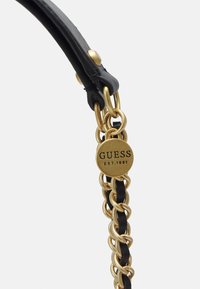 Cintura in pelle nera con dettagli a catena dorata intrecciata. Presenta un'etichetta tonda in oro incisa con "GUESS EST.1981." Texture liscia.