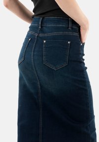 Denimskørt i mørkeblå med en tætsiddende silhuet, baglommer med sølvhardware og synlige stikninger langs sømmene.