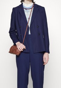 Personne portant un costume croisé bleu marine avec un pull léger bleu clair à motif et tenant un sac bandoulière marron.