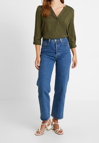 Blouse boutonnée vert olive avec un col en V, associée à un jean bleu moyen taille haute, droit, et des sandales blanches à brides.