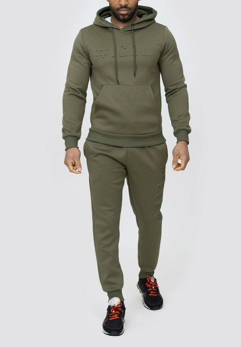 Ensemble de survêtement vert olive en tissu doux, comprenant un sweatshirt à capuche avec un logo texturé et un pantalon assorti avec des accents sur les côtés.