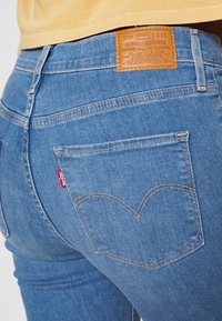 Jeans de mezclilla azul con un ajuste de talle medio, un parche de cuero marrón con el logo en la cintura y una etiqueta roja en el bolsillo trasero. Detalles de costura clásicos.