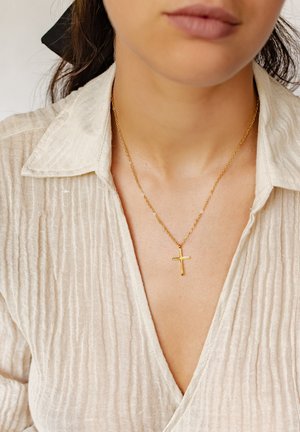Gold cross necklace on a delicate chain, worn over a textured, light-colored shirt with a deep V-neck and subtle vertical ribbing.