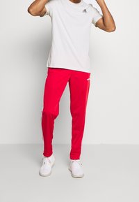 Czerwone spodnie sportowe z białymi paskami po bokach, wykonane z lekkiego materiału, w połączeniu z jasnoszarym T-shirtem i białymi trampkami.