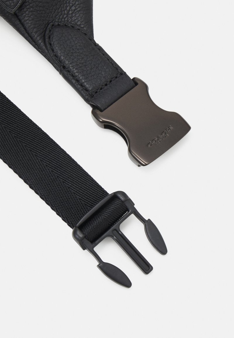 Zwarte leren band met een gestructureerde afwerking, voorzien van een donkere metalen gesp. Een uiteinde van de nylon band met een zwarte plastic sluiting is ook zichtbaar.