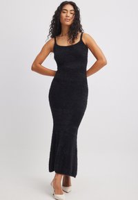 Black, sleeveless maxi dress made of soft, textured fabric, featuring a fitted silhouette and flared hemline. Polished light-colored heels.
