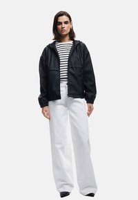 Zwarte waterdichte jas met capuchon, twee voorzakken, gedragen over een wit en marineblauw gestreept shirt, gecombineerd met wijde witte broeken.