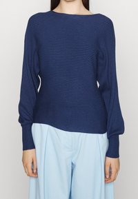 Granatowy sweter prążkowany z szerokim dekoltem w łódkę i długimi rękawami, zestawiony z jasnoniebieskimi spodniami szerokimi na nogawkach. Widoczna fakturowa tkanina.