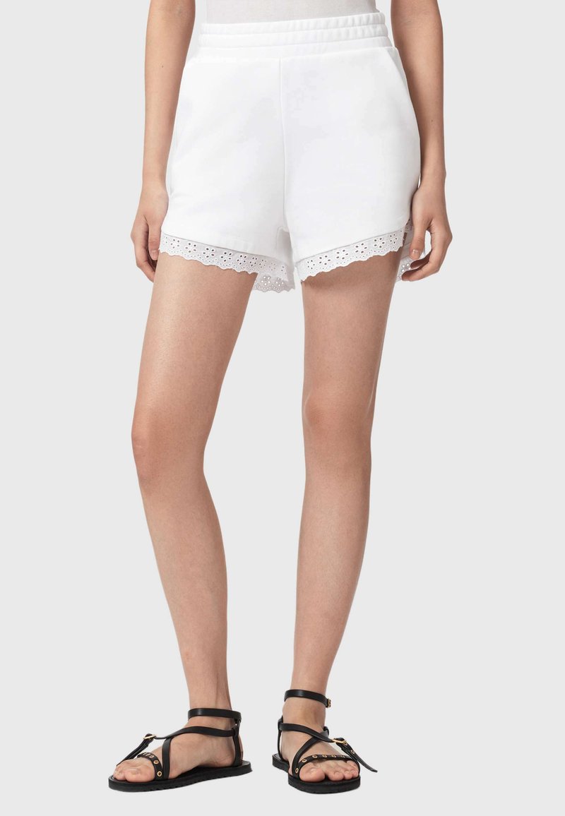 Woman wearing white high-waisted shorts with eyelet lace trim, paired with black flat sandals on a neutral background.