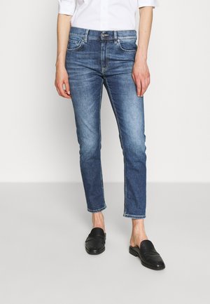 Osoba nosząca niebieskie dżinsy z wysokim stanem o dopasowanym kroju i czarne wsuwane mule, stojąca na jednolitym jasnym tle.