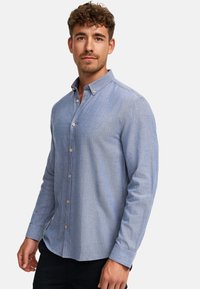 Lyseblå skjorte med knapper, lavet af tekstureret stof, med krave og lange ærmer med brune knapper.