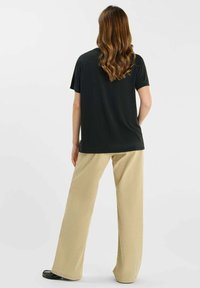 Vrouw met lang golvend haar, gekleed in een zwart shirt met korte mouwen, beige brede broek en zwarte schoenen, staand met de rug naar de camera tegen een effen achtergrond.