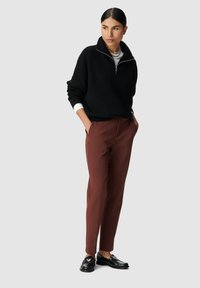 Donna che indossa un maglione nero a mezzo zip, una maglietta bianca, pantaloni marroni su misura, mocassini neri e una collana a catena, in piedi con le mani nelle tasche.