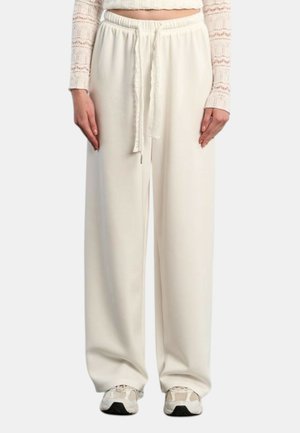 Femme portant un pantalon crème taille haute, jambes larges, avec un cordon à la taille, associé à des baskets blanches et un top court en dentelle à manches longues.