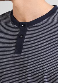 Chemise henley rayée bleu marine et blanche avec un col arrondi, deux boutons noirs et un tissu texturé près du cou.